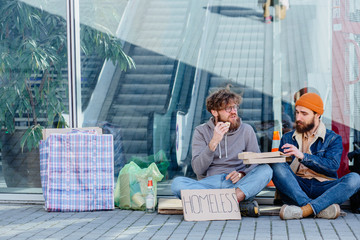 Homeless beggar and young handsome beard man listening to his story sitting together on the ground outdoors. Concept of human understanding