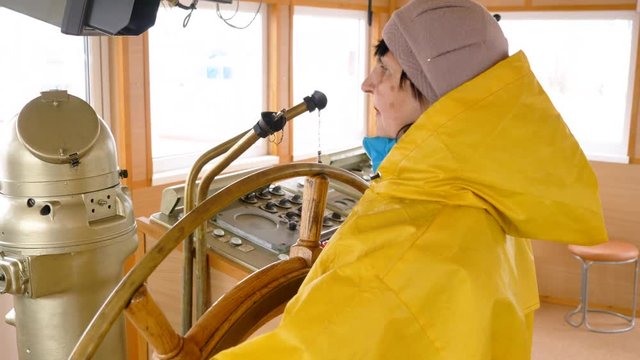 Senior woman in yellow raincoat rotates the steering wheel on the ship. She is in wheelhouse of the marine vessel.