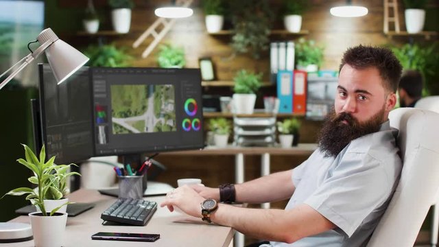 Bearded videographer working on dual screen setup in cozy loft eviroment. He looks at the camera ans smiles