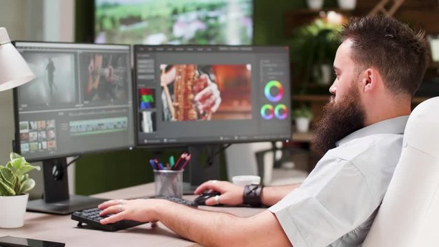 Bearded guy work as video editor or colorist in creative media agency. In the background - modern office with big TV displaying footage