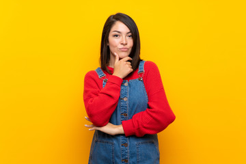 Young Mexican woman with overalls over yellow wall thinking