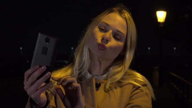 A young beautiful woman takes selfies with a smartphone in an urban area at night - streetlight in the background