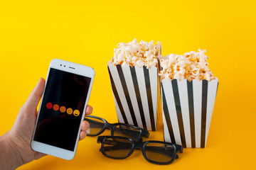 A hand is holding a smartphone in front of black-and-white paper boxes with popcorn and 3D glasses