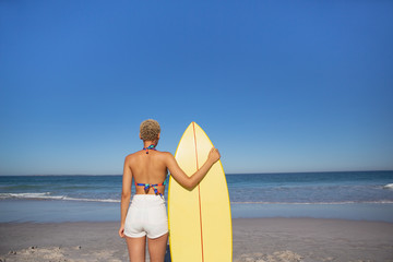 Woman in bikini standing with surfboard on beach in the sunshine