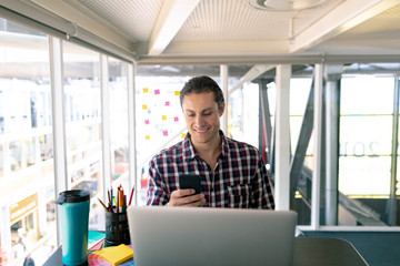 Fototapeta premium Male graphic designer using mobile phone while working on laptop at desk