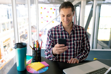 Male graphic designer using mobile phone while working on laptop at desk