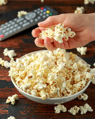 Hand takes salty popcorn from the bowl and remote control on wooden table