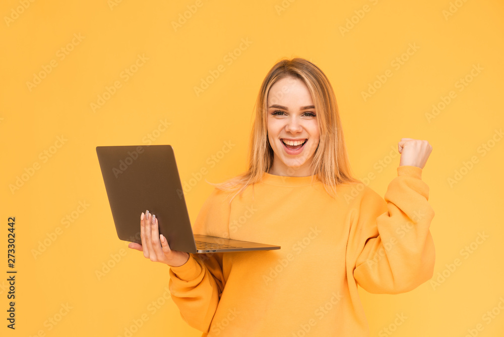 Wall mural blonde in yellow clothes rejoices in victory with a laptop in their hands. portrait of a happy girl 