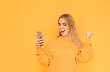 A smiling blond girl uses a smartphone in bright clothes and rejoices on a yellow background. Portrait of a happy girl looking at the screen of a smartphone and rejoices in victory, isolated.
