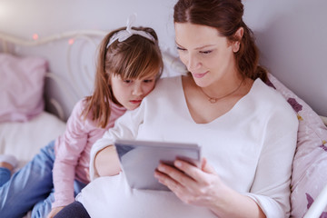 Beautiful Caucasian pregnant woman sitting on sofa with her cute daughter and using tablet.