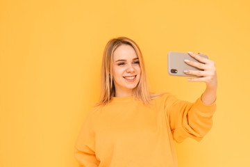 Attractive young lady in bright clothes makes selfie on a yellow background, posing for the camera smartphone, smiling and looking at the phone. Isolated. Copy space