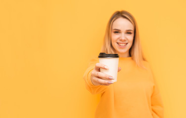 Smiling blonde in an orange sweater shows a cup of coffee in the camera on a yellow background, looking into the camera.Ppaper cup of coffee in the hands of a positive girl. Focus on a cup of coffee - Powered by Adobe
