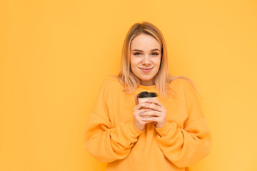 Girl in an orange sweater stands on a yellow background with a cup of coffee in her hand, looks at...