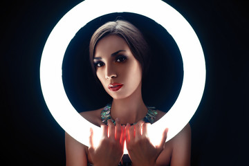 A beautiful young Caucasian girl with a beautiful make-up looks out from behind a circular lamp for...
