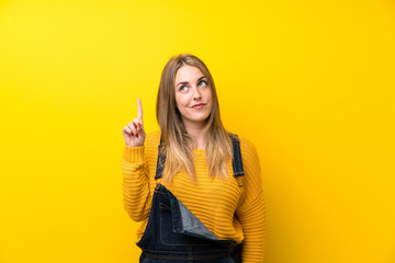 Woman with overalls over isolated yellow wall pointing with the index finger a great idea