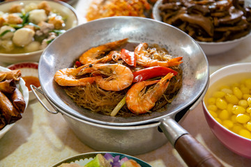 Casseroled prawns with Glass Noodels in Small Iron Pan with Fish, Fried Spring Rolls, Dim Sum, Seafood, Roasted Duck and Meatball Background on the Table.