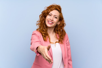 Redhead woman in suit over isolated blue wall shaking hands for closing a good deal