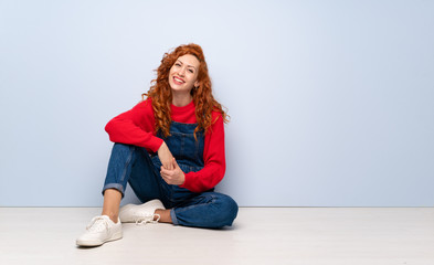 Redhead woman with overalls sitting on the floor keeping the arms crossed in frontal position