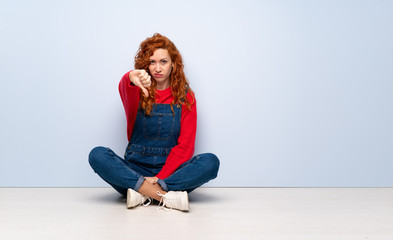 Redhead woman with overalls sitting on the floor showing thumb down with negative expression