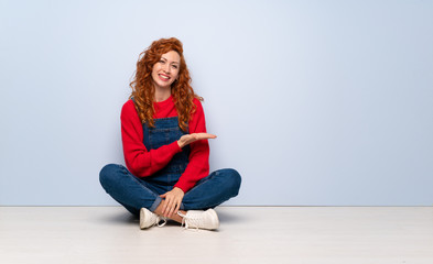 Redhead woman with overalls sitting on the floor presenting an idea while looking smiling towards