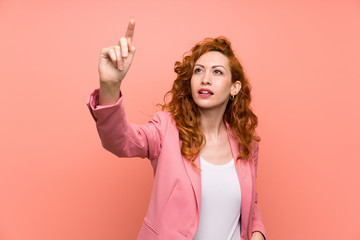 Redhead woman in suit over isolated pink wall touching on transparent screen