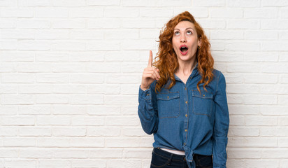 Redhead woman over white brick wall pointing up and surprised