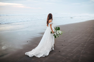 Fototapeta na wymiar Bride on a beach in the blue water with a bouquet in hands on sunset
