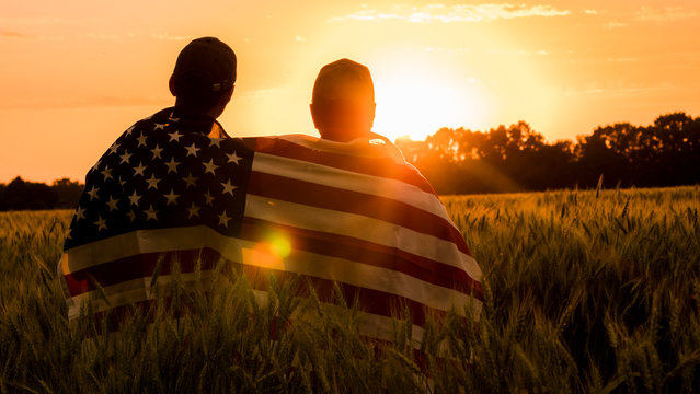 A man and his son admire the sunset over a field of wheat, wrapped in the flag of the USA