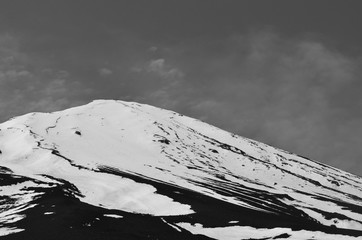 The peak of a mountain is seen against a cloudy sky. The mountain is covered in snow. The photograph is in black and white.