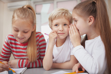 Gossip, bullying, secrets concept. Adorable blond haired little girl smiling, looking away thoughtfully, while her friend whispering to her ear. Elementary school pupils chatting quietly during art cl