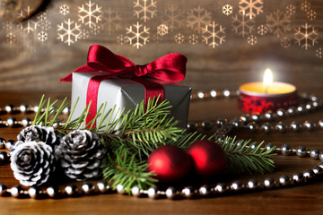 Christmas composition: a gift wrapped with red ribbon, silver cones, red baubles and a fir tree branch - burning candle in background, snow flakes above