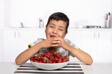 Shot of a smiling boy sitting in the kitchen eating strawberries with closed  eyes from pleasure. © rozaivn58