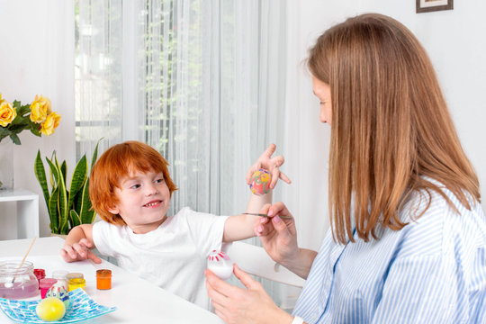 Little redhead boy with a nanny or mother or teacher sit at the table and paint with colors.