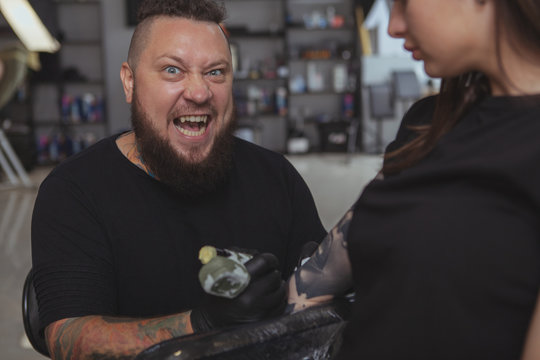 Bearded Expressive Tattoo Artist Growling, Looking Scary  While Tattooing Female Client. Cropped Shot Of Unrecognizable Woman Getting Tattooed By Crazy Tattooer