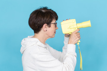 Side view of a middle-aged woman with a short haircut in a sweater and glasses holding a yellow vintage movie camera. Concept of video shooting