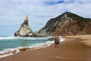 Happy man and woman couple walking on a deserted beach with sand and rocks. Rear view.