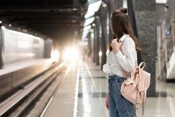 young european girl in jeans and shirt with backpack is waiting for the train at the metro station. Background in motion blur to convey a vibrant atmosphere. This is a snapshot idea.