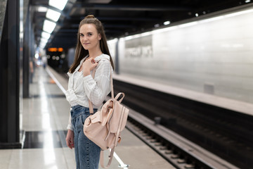 European girl with a backpack standing at the subway station and waiting for the train. The train is out of focus to convey the effect of the reality of the movement. The main object is a girl.