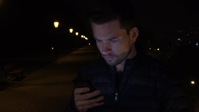 A young handsome man works on a smartphone in an urban area at night - a line of streetlights in the background