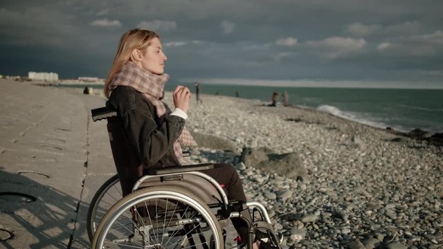 Young blonde woman with paralysis of legs is sitting in wheelchair, viewing sea