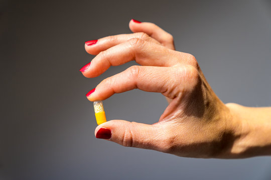 Pretty female hand with red fingernails holding a single medicine pill between fingers presenting healthy supplement with vitamins on grey background