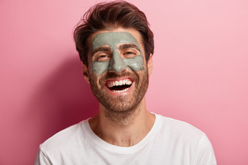 Joyful delighted man has clay mask on face, enjoys spa treatments, has broad smile, being in high...