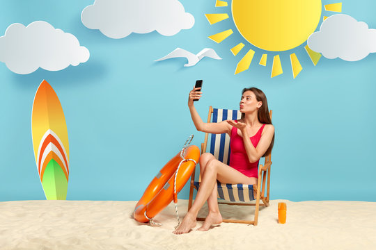 Lovely female model blows air kiss at smartphone camera, makes selfie, poses at beach chair, wears red bikini, sunbathes at coast, expresses love to boyfriend on distance, being in resort place