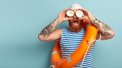 Surprised male lifeguard keeps two coconuts, has tattooed arms, wears striped sailor vest, carries orange lifering, cares about safety on water, isolated over blue wall. Boatswain with swim equipment