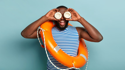 Funny male life preserver carries two coconuts near eyes, wears striped vest, lifebuoy with rope on...