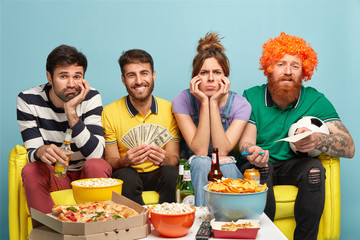 Sad company of friends feel bored as watch not interesting match on television, surrounded with different delicious snacks, happy guy holds money, makes bet on game, pose at comfortbale sofa