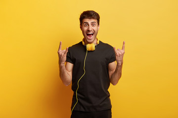 I like heavy metal! Joyful young male being crazy, makes horn gesture with both hands, listens favourite music in headphones, keeps mouth opened, wears casual black t shirt, stands over yellow wall