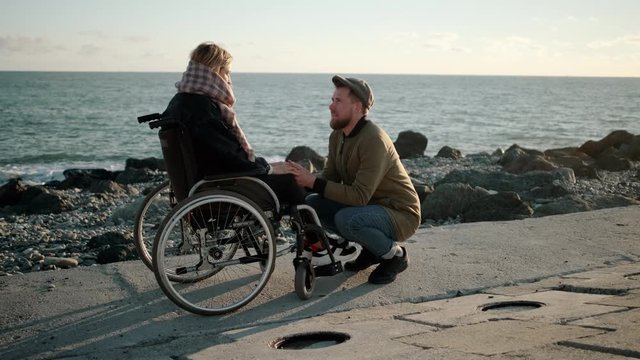 Man is holding hands of sick young woman in wheelchair and talking with her