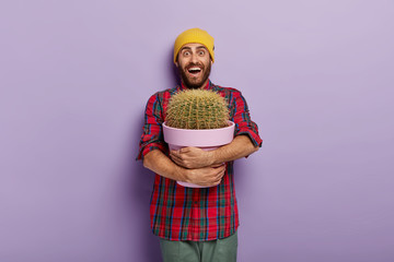 Happy Caucasian man embraces pot with big cactus, being plant lover, receives indoor plant as present, cannot believe his eyes, wears yellow hat and checkered shirt, poses against purple studio wall