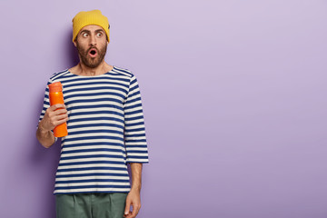Studio shot of impressed student drinks hot coffee from thermos bottle during break, focused aside with astonishment, dressed in stylish apparel, models against purple wall, copy space for text